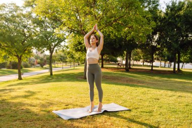 Atletik kıyafetleri ve kablosuz kulaklıkları olan güzel bir kadın parkta yoga yapıyor. Atletik kadın güneşli bir günün tadını çıkarıyor, meditasyon yapıyor ya da dışarıda egzersiz yapıyor. Egzersiz ve rahatlama kavramları.