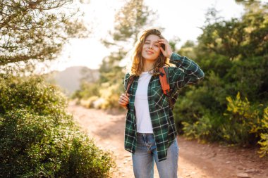 Sırt çantalı genç bir kadın yeşil bitkilerle çevrili dağ yolunda yürüyor. Dişi yürüyüşçü gün batımında dağ manzarasını ve doğayı sever. Konseptler: macera, rahatlama, aktif yaşam tarzı.