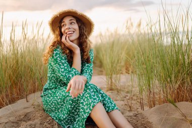 Hasır şapkalı ve çantalı mutlu bir kadın gün batımında kumsalda yürümekten zevk alıyor. Güneşli bir günde güzel bir kadın sahilde poz verir. Güzellik, hafta sonları ve doğa kavramı..