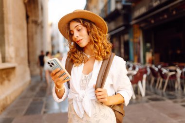 Şapkalı şık bir kadın ve elinde telefonla şehrin güneşli caddelerinde sırt çantasıyla geziyor. Telefonu olan güzel bir turist gün batımında mimariye hayran kalır. Turizm ve teknoloji kavramı.