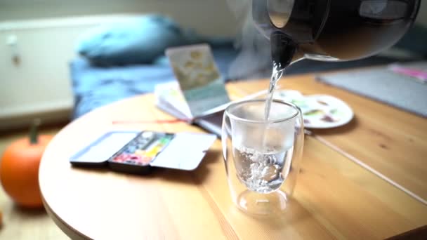 Mujer vierte agua caliente de la tetera negra en un vaso cerca de la paleta con pinturas — Vídeos de Stock