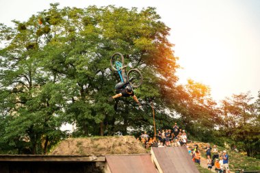 Bisikletli bir sürücü yeşil ağaçların yanında gösteri yapıyor ve insanlar gün batımında.