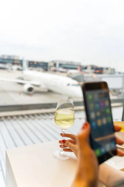 Female hand holding smart phone and glass of wine making picture near airplane