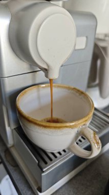 Kahve makinesinden sıcak bir mutfak ortamında seramik bir fincana taze espresso dökülüyor. Görüntü sabah rutinini, kafein artışını ve kahve yapma sanatını temsil ediyor.