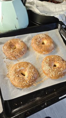 Mutfak ortamında parşömen kağıdının üzerinde duran taze susamlı simit. Görüntü ev yapımı pişirme, konforlu yiyecekler ve evde basit ekmek yapma zevkini temsil ediyor.