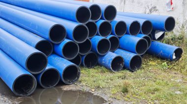 Duvarın yanındaki ıslak çimenlerin üzerinde duran büyük mavi plastik borular yığını. Görüntü inşaat, sanayi ve altyapı gelişimini temsil ediyor