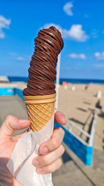 Waffle külahında uzun bir çikolatalı dondurmayı güneşli bir plaj arka planına karşı tutarak. Bu resim neşe, yaz ziyafeti ve kaygısızlığı iletir.