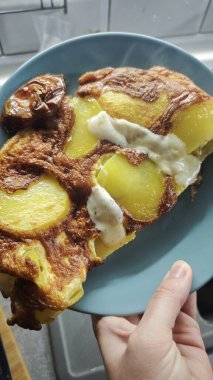 Eritilmiş peynirli patates omleti dilimi mavi tabakta servis edilir. Bu resim ev yapımı konforlu yemek ve sade kırsal yemek kavramını aktarıyor.