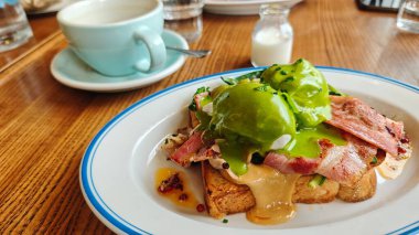 Kızarmış ekmeğin üzerinde domuz pastırmalı Benedict yumurtaları yeşil hollandaise ile kaplanır ve bir fincan kahvenin yanında servis edilir. Bu görüntü, doyurucu brunch ve hoşgörülü kafe yemeği kavramını aktarıyor.