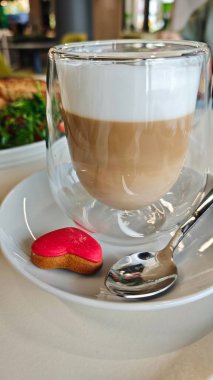 Süt köpüklü bir bardak sütlü latte, kalp şeklinde kurabiye ve kaşık şeklinde kaşık. Kahve molası sahnesi aşkı, rahatlığı ve rahatlığı simgeliyor.