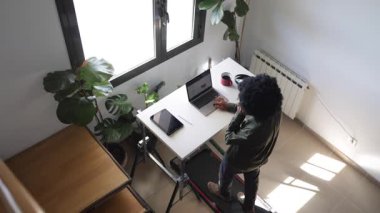 Bilgisayarlı, telefonlu, kulaklıklı, küçük bir koşu bandının üzerinde telefonda konuşan Afro-Amerikalı genç bir adam.