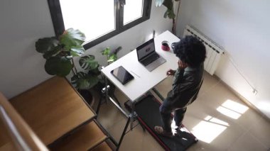 Bilgisayarlı, telefonlu, kulaklıklı, küçük bir koşu bandının üzerinde telefonda konuşan Afro-Amerikalı genç bir adam.