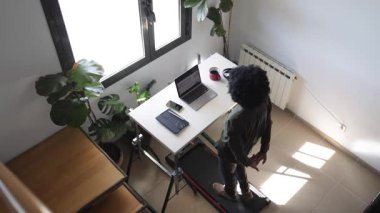 Bilgisayarlı, telefonlu, kulaklıklı genç Afro-Amerikalı adam küçük bir koşu bandının üzerinde masanın üstünde.