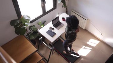 Bilgisayarlı, telefonlu, kulaklıklı genç Afro-Amerikalı adam küçük bir koşu bandının üzerinde masanın üstünde.