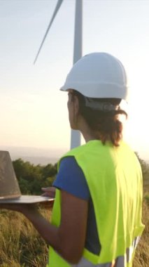 Güvenli Yelek 'te bir kadın, türbinlerle dolu bir alanda dururken dizüstü bilgisayar kullanıyor.  