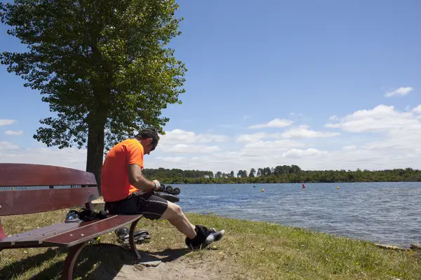 Парень, чинивший коньки, летом сидит на скамейке у озера. — стоковое фото