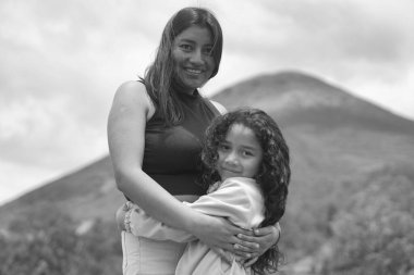 Anne ve kızı bir arada dururken gülümsüyor, arkalarında dağ manzarası ile sevgi dolu bir kucaklaşmayı paylaşıyorlar, aile bağını ve çocukluk mutluluğunu temsil ediyorlar.