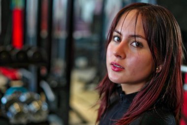Kızıl saçlı ve parlak tenli genç bir kadın kameraya bakıyor. Spor salonundaki fitness eğitimi sırasında odaklanmış bir kararlılık gösteriyor.
