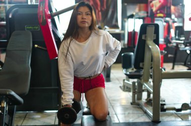 Genç Asyalı bir kadın spor salonundaki bir bankta halterle odaklanmış güç çalışması yapıyor. Kararlılığını, kas gelişimini ve aktif sağlıklı yaşam tarzını gösteriyor.