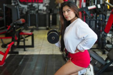 Asyalı bir kadın dambılı kaldırıyor, fitness merkezinde egzersiz yapıyor, kas geliştirmek ve sağlıklı bir yaşam tarzı sürdürmek için biseps buklelerine odaklanıyor.