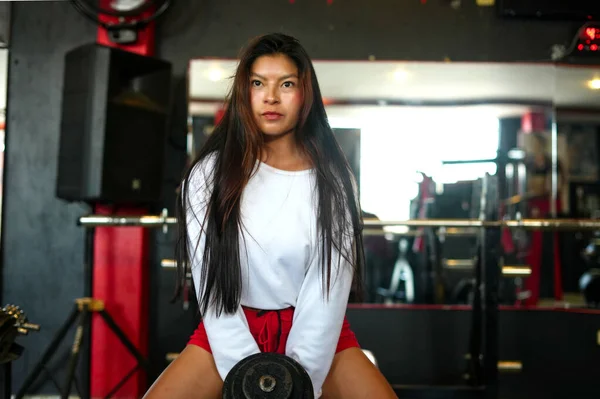 Ciddi bir genç kadın, spor salonunda tek bir ağır dambılı kaldırıp sağlıklı ve aktif yaşam tarzının bir parçası olarak egzersizine odaklanıyor.