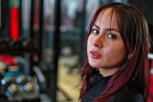 Kızıl saçlı ve parlak tenli genç bir kadın kameraya bakıyor. Spor salonundaki fitness eğitimi sırasında odaklanmış bir kararlılık gösteriyor.
