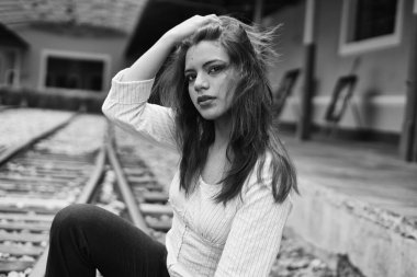 Genç bir kadın, siyah-beyaz bir şehir portresi içinde demiryolunda oturuyor, uzun saçları elinde, düşünceli ve şık, grunge sokak modasını düşünceli bir ifadeyle harmanlıyor.