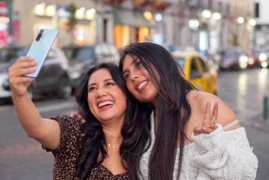 İki neşeli bayan arkadaş gülümsüyor ve akıllı bir telefondan bir gece şehir selfie 'si için poz veriyorlar. Bir tanesi birlikte neşeli ve samimi bir anı paylaşırken barış işareti gösteriyor.