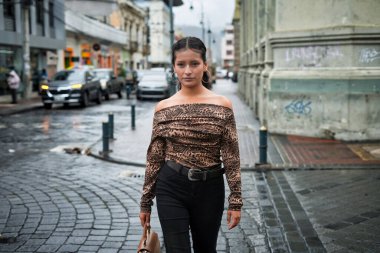 Genç Latin Amerikalı kadın, ıslak kaldırım taşı şehir caddesinde, omuzları açık, siyah pantolon ve el çantası içinde, şehir mimarisi ve grafitisine karşı poz veriyor.