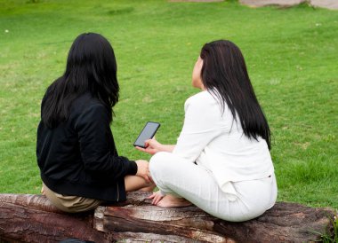 Parktaki bir kütükte oturan iki kadın, biri akıllı telefonu tutarken sohbet ediyorlar, yeşil bir ortamda iletişim ve iletişim anı yaratıyorlar.