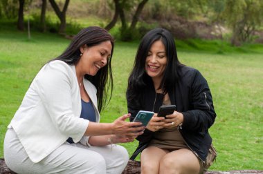 İki neşeli kadın parkta birlikte oturuyor, birbirlerinin arkadaşlığından keyif alıyor ve cep telefonlarıyla eğlenceli bir şey paylaşıyorlar, teknoloji aracılığıyla iletişim kuruyorlar.