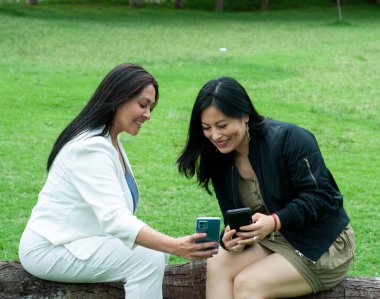 İki neşeli bayan arkadaş açık havada oturuyorlar, akıllı telefondan içeriklerini paylaşıyorlar ve parkta birlikte gülüyorlar, boş zamanlarının ve bağlantılarının tadını çıkarıyorlar.