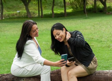 Yeşil bir parkta kütüğün üzerinde oturan iki kadın, cep telefonunda gülümseyen ve içeriği paylaşan, açık hava teknolojisiyle dostça iletişim ve bağlantının keyfini çıkaran iki kadın.