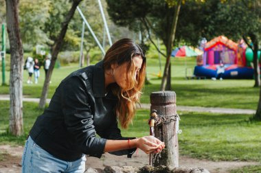 Genç bir kadın, güneşli, yeşil bir yaz gününde serinleyip, köy parkı çeşmesinin temiz sularında ellerini yıkıyor, susuzluğun ve açık havada dinlenmenin keyfini çıkarıyor.