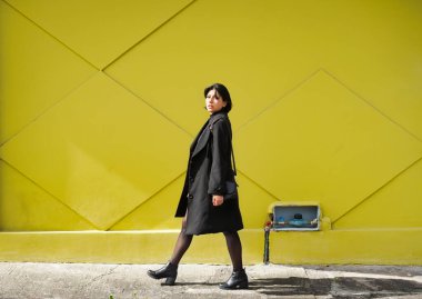 Siyah paltolu ve çizmeli bir kadın, güneşli bir şehir caddesinde, geometrik desenli canlı sarı bir duvara karşı güvenle yürüyor, modern sokak tarzını ifade ediyor.