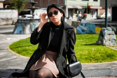 Güneş gözlüklü, siyah ceketli ve ekose etekli bir kadın şehir parkında alçak bir duvarda oturuyor, bir çanta tutuyor ve yukarı bakıyor, modern tarzla şehir hayatını birleştiriyor.