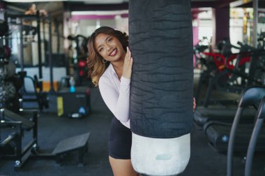 Kendine güvenen bir gülümsemesi olan genç bir kadın modern bir spor salonunda ağır bir kum torbasının arkasından zirve yapıyor. Formunu, sağlığını ve egzersize karşı olumlu bir tutum sergiliyor.