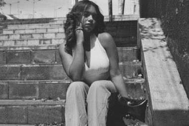 Bir şehirde beton basamaklarda oturan genç bir kadın, siyah-beyaz, düşünceli ve yalnız, derin derin düşüncelere dalmış, ince düşüncelere dalmış, yüzünde güneş ışığı ve gölgeler vardı.