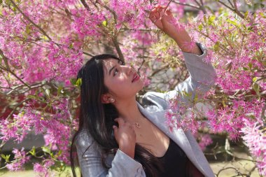 Kadın dışarıda duruyor, narin pembe Loropetalum çiçeklerine uzanıyor, doğanın güzelliğinin ve bahar güneşinin sıcaklığının tadını çıkarıyor.