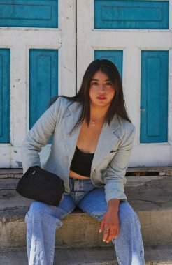 Basamaklarda oturan kendine güvenen genç bir kadın şık bir spor ceket ve kot pantolon giymiş bir portre için poz veriyor, canlı mavi yıpranmış bir kapı ile renkli bir şehir zemini yaratıyor.