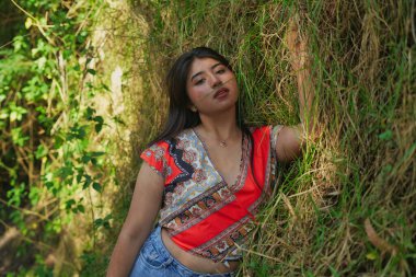 Genç bir kadın uzun yeşil çimenlerde uzanıyor, ılık yaz güneşinin ve temiz havanın tadını çıkarıyor, huzurlu bir çayır ortamında doğayla iç içe geçiyor, kameraya bakıyor.