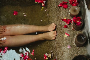 Kırmızı gül yapraklarıyla dolu sıcak bir küvette bacaklarını ve ayaklarını ıslatan bir kadın, yumuşak mum ışığında, spa benzeri bir ortamda rahatlık ve kendine bakım buluyor.