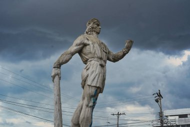 Taştan heykel, elinde bir sopa ve gemi tutan yerli bir savaşçının dramatik bulutlu gökyüzüne gururla karşı duruşu tarihi cesaret ve kültürel dayanıklılığı simgeliyor.