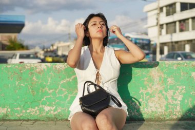 Genç bir kadın dokulu yeşil bir duvarda oturuyor, koyu renk saçlarını düzeltiyor, beyaz bir bluz ve şort giyiyor, canlı bir şehir sokak ortamında güneş ışığıyla dolu siyah bir çanta tutuyor.