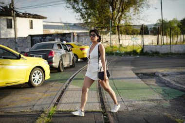 Genç bir kadın şık beyaz bir yaz kıyafeti ve güneş gözlükleriyle işlek bir caddeden güvenli bir şekilde geçiyor. Arabalar beklerken ve gün ışığı şehir yaşamını aydınlatırken amaçla yürüyor.