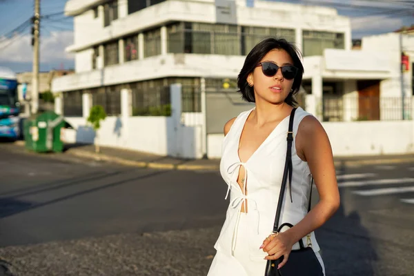 Şık beyaz elbiseli ve güneş gözlüklü genç bir kadın altın saatinde güneşli bir caddede güvenli bir şekilde yürüyor, modern sokak stilini, yaz rahatlığını ve bağımsız zarafeti somutlaştırıyor.