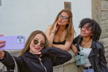 Üç farklı gülümseyen genç kadın cep telefonuyla kendi portresini çekiyorlar, şehirde bir anı paylaşırken arkadaşlığı ve eğlenceyi kucaklıyorlar.