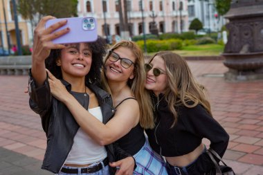 Şehirli bir meydanın keyfini çıkarırken üç farklı genç kadın neşeli bir şekilde gülümsüyor, mutlu anıları bir araya getiriyor ve akıllı telefonlu bir selfie ile birleştiriyor.