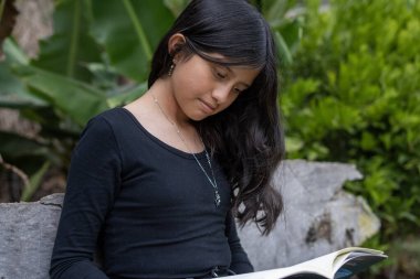Koyu renk saçlı genç bir kız, bir kitaba dalmış, yeşil bir bahçede, taştan bir bankta otururken konsantre oluyor, doğada huzurlu edebiyatın tadını çıkarıyor.