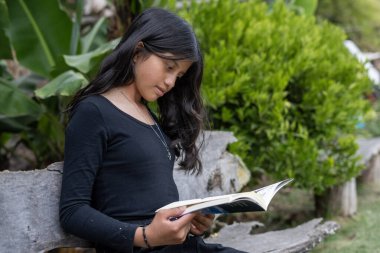Odaklanma ve konsantrasyonla kitap okuyan genç bir kız açık havada ahşap bir bankta rahatça otururken eğitimi, edebiyatı ve doğanın huzurlu ortamını kucaklıyor.